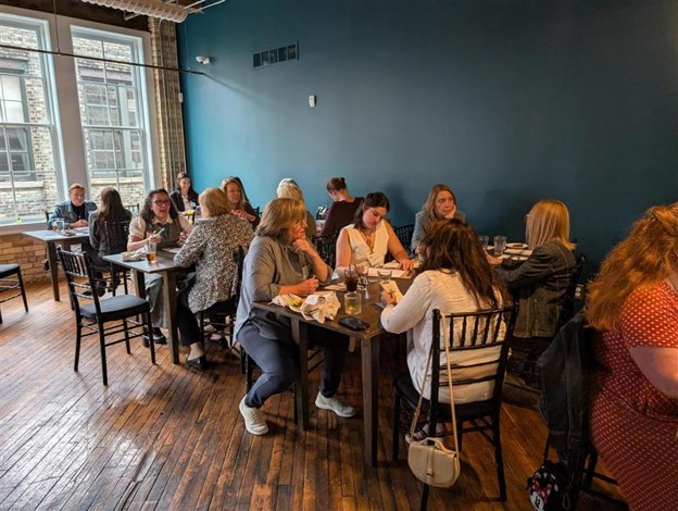 Photograph of a group of people dining and networking in a restaurant with wooden floors and large windows providing natural light. The setting features dark blue walls, black chairs, and tables with food and drinks, capturing a casual social gathering atmosphere.