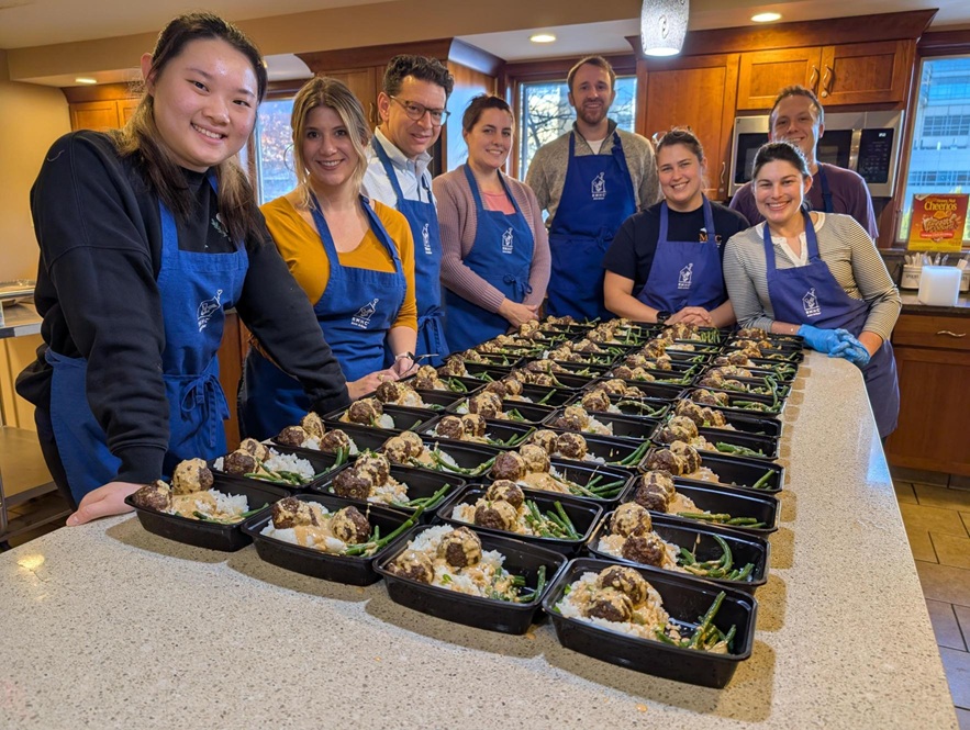 Photograph of MVC Coordinating Center team volunteering at RMHCAA wearing blue aprons standing behind a kitchen counter filled with multiple meal prep containers of food.